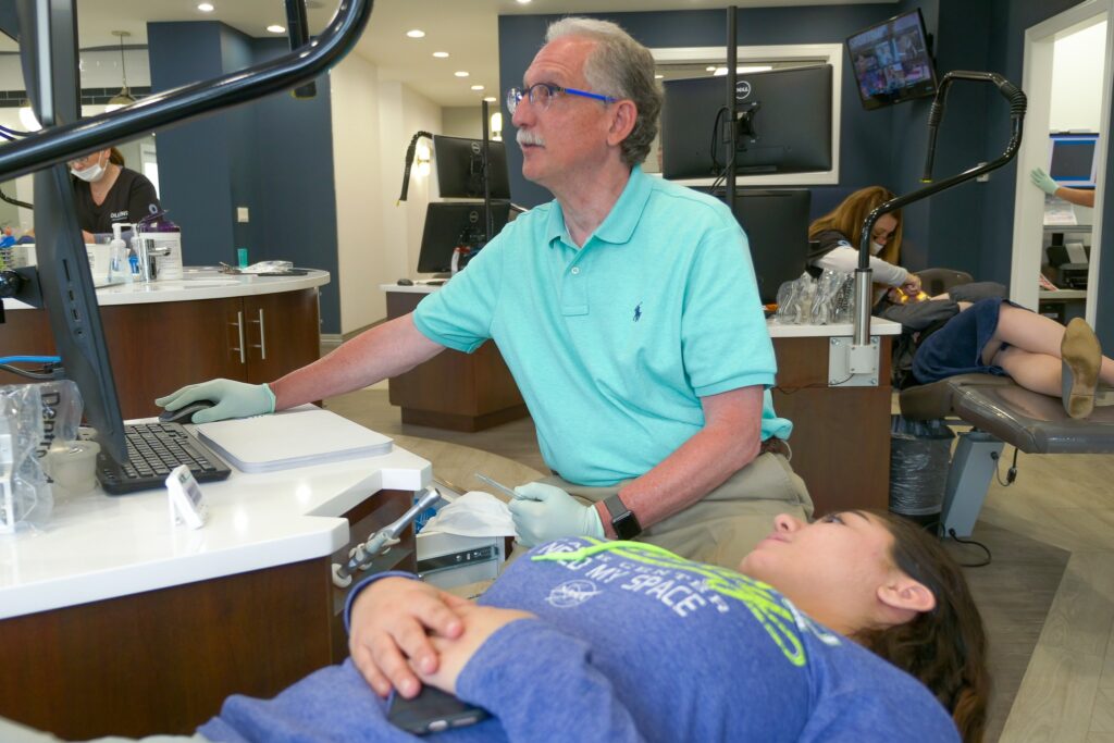 Dr. Bruce Ollins in a teal polo checks a young patient’s braces at a treatment chair in the Ollins Orthodontics office in Nutley, NJ